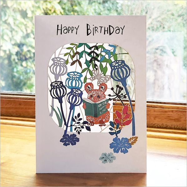Birthday card with a fox reading a book among flowers on a wooden surface.